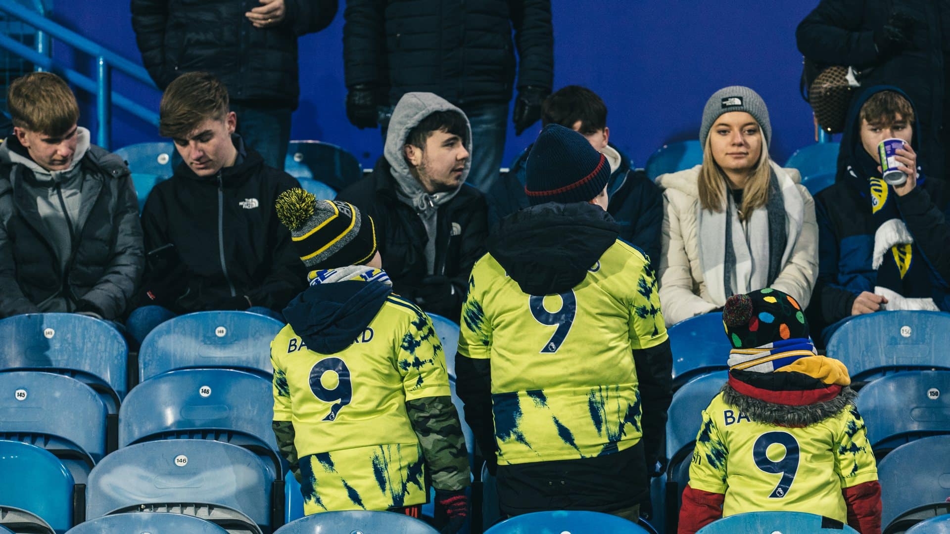 Three young Leeds fans turning their backs and showing three identical 'Bamford 9's on the back of their blue cheese away shirts