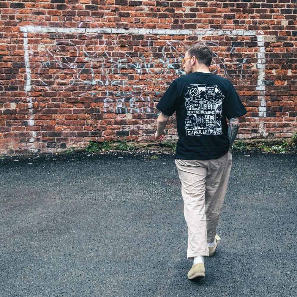 A photograph of the back of our new TSB crowd t-shirt, featuring an illustration by Josh Parkin of a stand at Elland Road filled with flags — Bielsa is God, Norman Bites Yer Legs, Keep Fighting, Vile Animals and other favourites