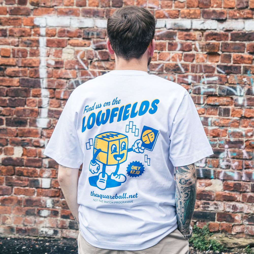 A photograph of the back of our new TSB mascot t-shirt, featuring a Square Ball with arms and legs selling an issue of the mag with our new funky Smiley badge-inspired logo, below the words 'Find us on the Lowfields'