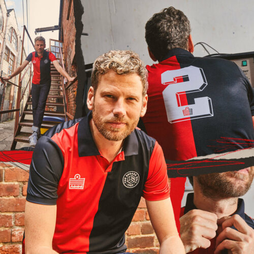Simon Rix from Kaiser Chiefs wearing the away edition of our TSB x Admiral collab football shirt, which is a half-black half red tribute to Marcelo Bielsa's Newell's Old Boys, with a button down collar, TSB and Admiral logos on the chest and a number 2 on the back