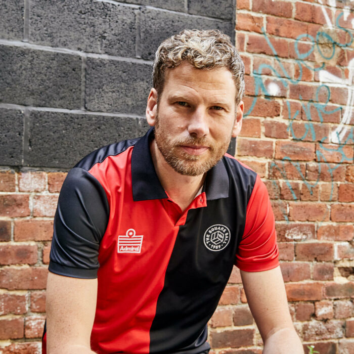 Simon Rix from Kaiser Chiefs wearing the away edition of our TSB x Admiral collab football shirt, which is a half-black half red tribute to Marcelo Bielsa's Newell's Old Boys, with a button down collar, TSB and Admiral logos on the chest and a number 2 on the back