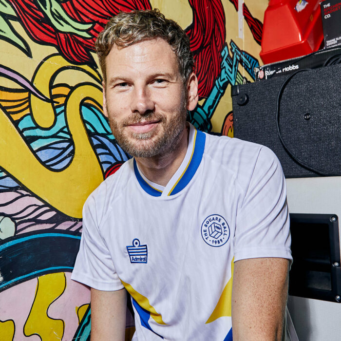 Simon Rix from Kaiser Chiefs wearing our TSB x Admiral collab football shirt, which is white with a blue v-neck collar, blue and yellow chest flashes, and the TSB and Admiral logos on the chest