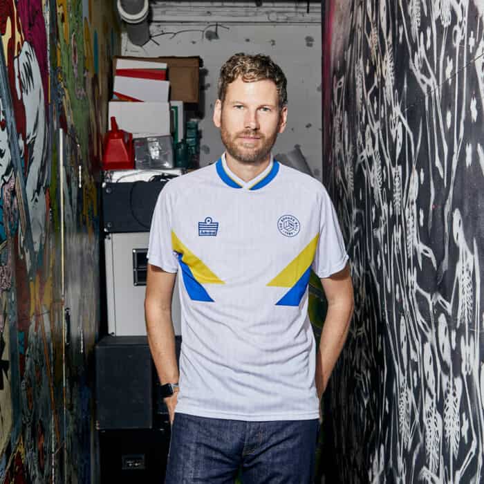 Simon Rix from Kaiser Chiefs wearing our TSB x Admiral collab football shirt, which is white with a blue v-neck collar, blue and yellow chest flashes, and the TSB and Admiral logos on the chest
