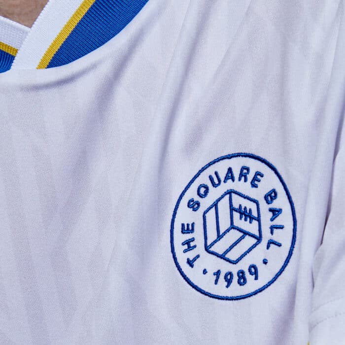 Simon Rix from Kaiser Chiefs wearing our TSB x Admiral collab football shirt, which is white with a blue v-neck collar, blue and yellow chest flashes, and the TSB and Admiral logos on the chest — this is a close up of the TSB badge