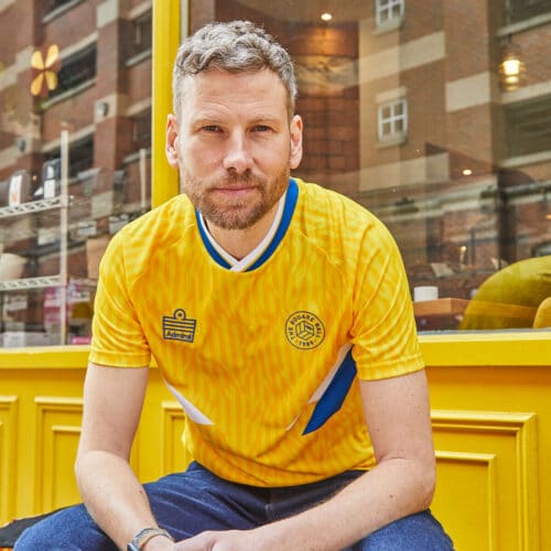 A photo of Simon Rix from Kaiser Chiefs outside Laynes Espresso in Leeds wearing our TSB x Admiral collab football shirt, which is yellow with a blue and white v-neck collar, white and blue chest flashes, and has the TSB and Admiral logos on the chest
