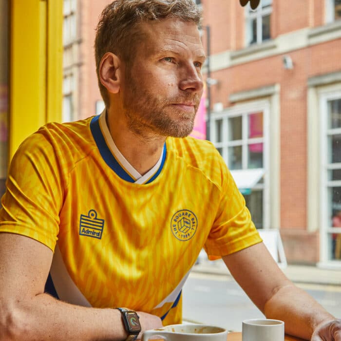 A photo of Simon Rix from Kaiser Chiefs inside Laynes Espresso in Leeds wearing our TSB x Admiral collab football shirt, which is yellow with a blue and white v-neck collar, white and blue chest flashes, and has the TSB and Admiral logos on the chest