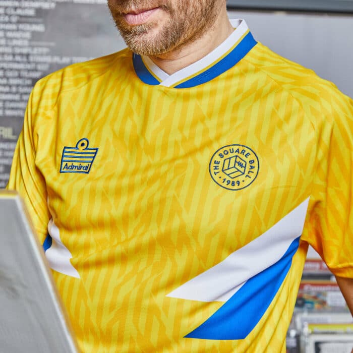 A close up photo of our TSB x Admiral collab football shirt, which is yellow with a blue and white v-neck collar, white and blue chest flashes, and has the TSB and Admiral logos on the chest