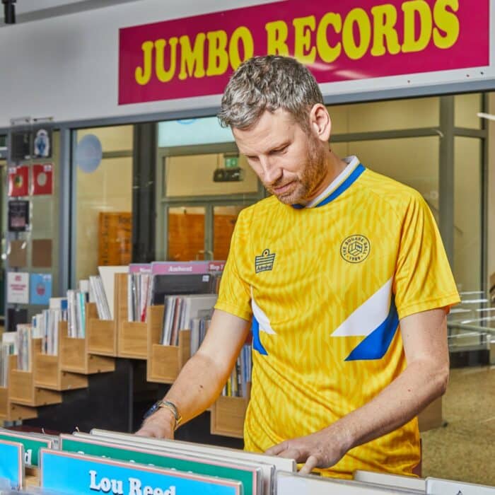 A photo of Simon Rix looking through the racks in Jumbo Records in Leeds wearing our TSB x Admiral collab football shirt, which is yellow with a blue and white v-neck collar, white and blue chest flashes, and has the TSB and Admiral logos on the chest