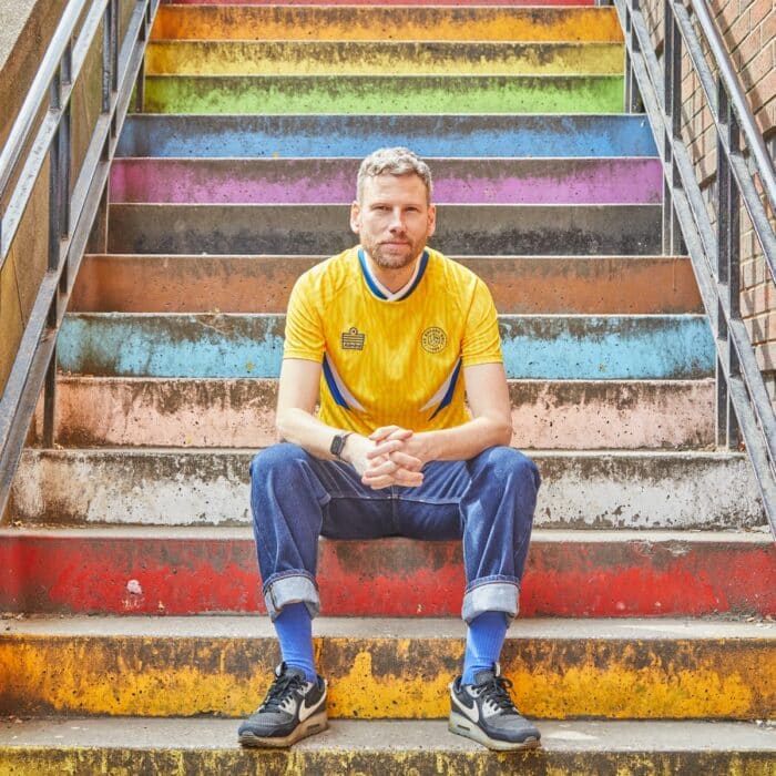 A photo of Simon Rix from Kaiser Chiefs sitting on the steps behind the Cockpit in Leeds wearing our TSB x Admiral collab football shirt, which is yellow with a blue and white v-neck collar, white and blue chest flashes, and has the TSB and Admiral logos on the chest