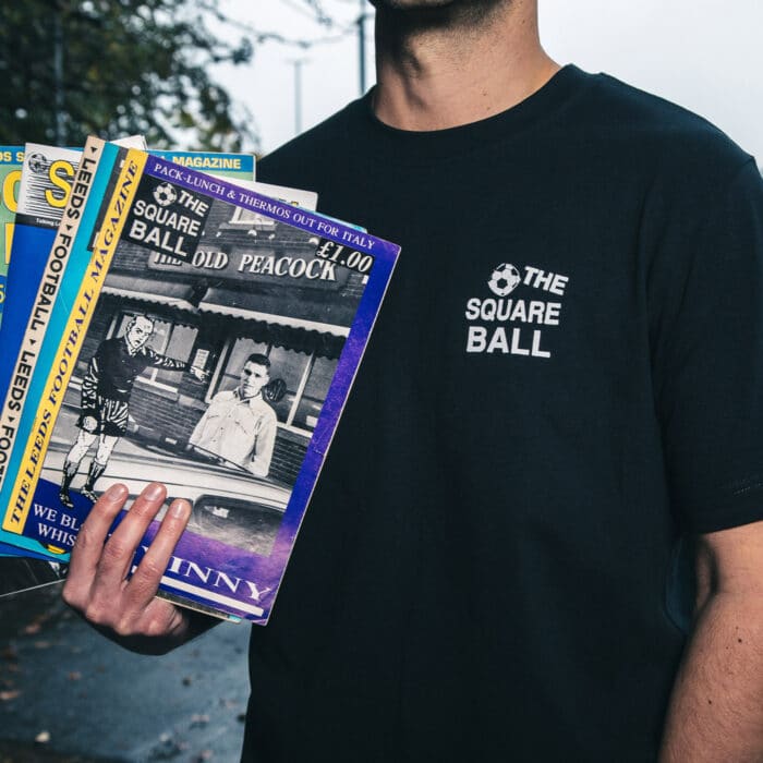 A photo of Michael wearing our retro logo t-shirt, showing a backprint featuring logos and graphics from throughout our history, and a front pocket print of the logo from the very first issue. He's holding some old copies of the magazine in the Lowfields Tunnel