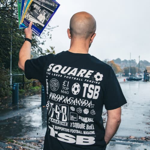 A photo of Michael wearing our retro logo t-shirt, showing a backprint featuring logos and graphics from throughout our history, and a front pocket print of the logo from the very first issue. He's holding some old copies of the magazine in the Lowfields Tunnel