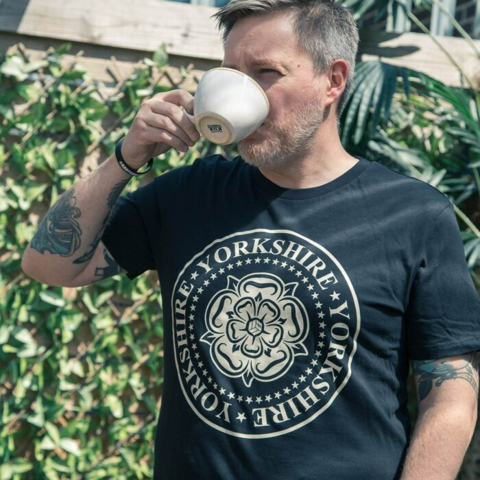 The TSB Yorkshire t-shirt, featuring a rose and icon design, being worn by a very handsome man who is drinking tea