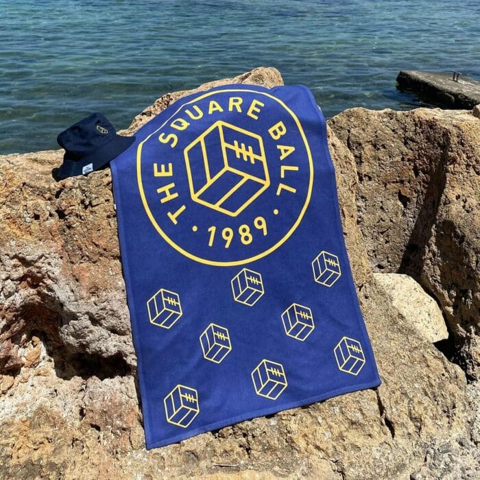 A blue TSB beach towel featuring our logo and icon in yellow, resting on some rocks by the sea