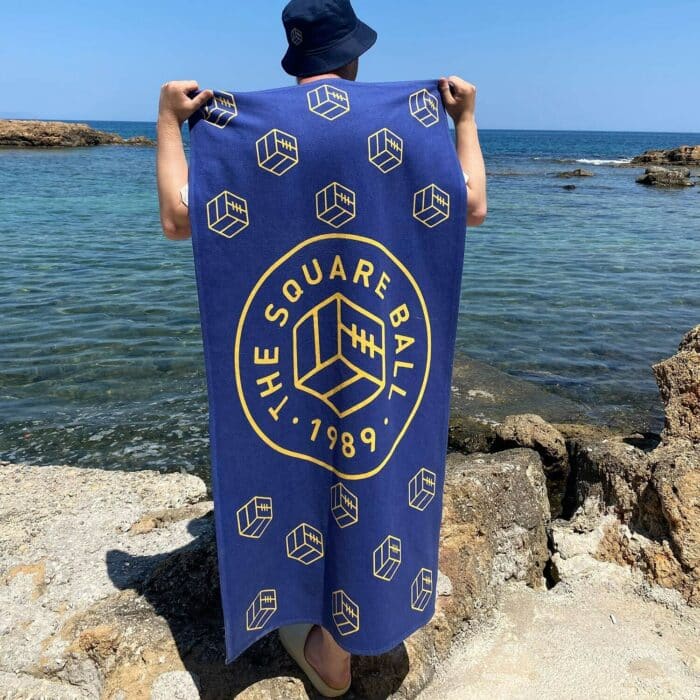 A blue TSB beach towel featuring our logo and icon in yellow being held up by a holidaymaker by the sea