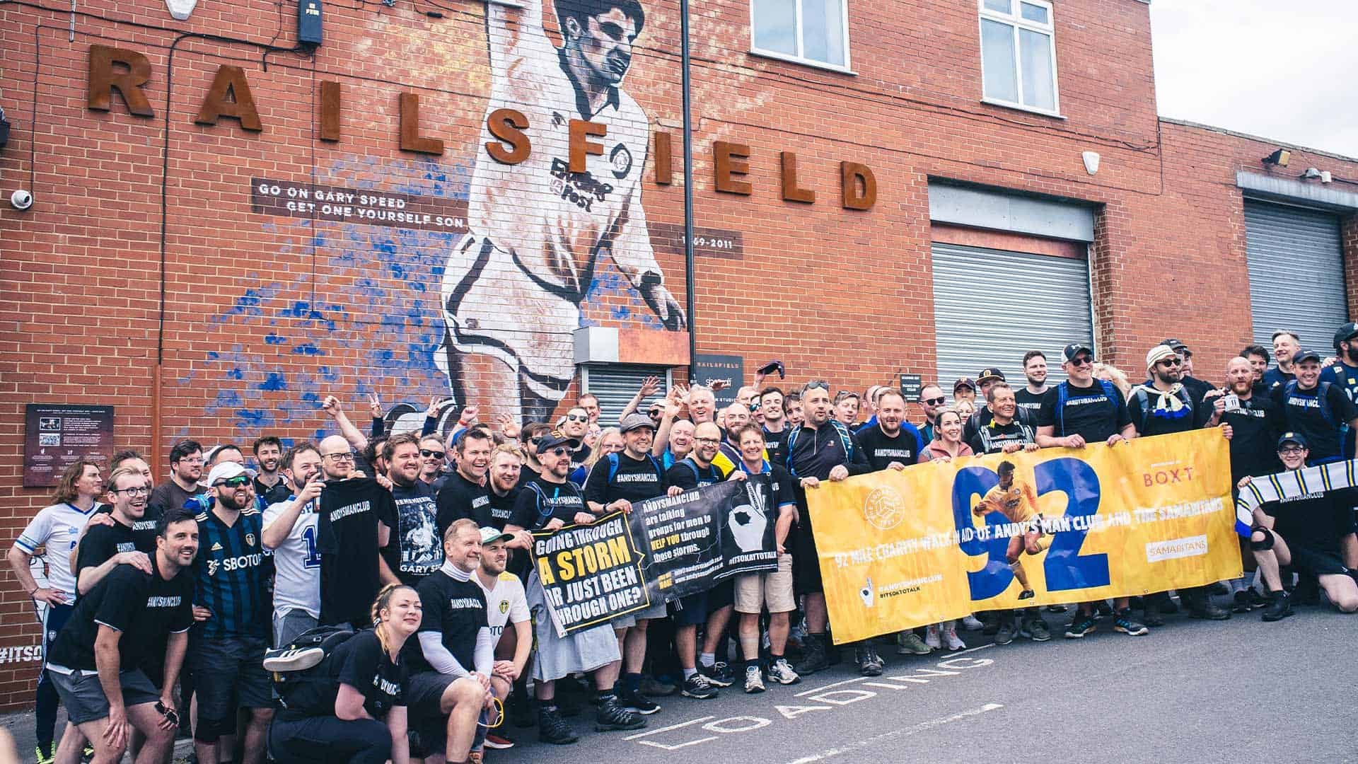 100 tired Leeds fans posing in front of the Gary Speed mural, holding a banner of Gary Speed and another for Andy's Man Club, around 90 miles into their 92 mile walk