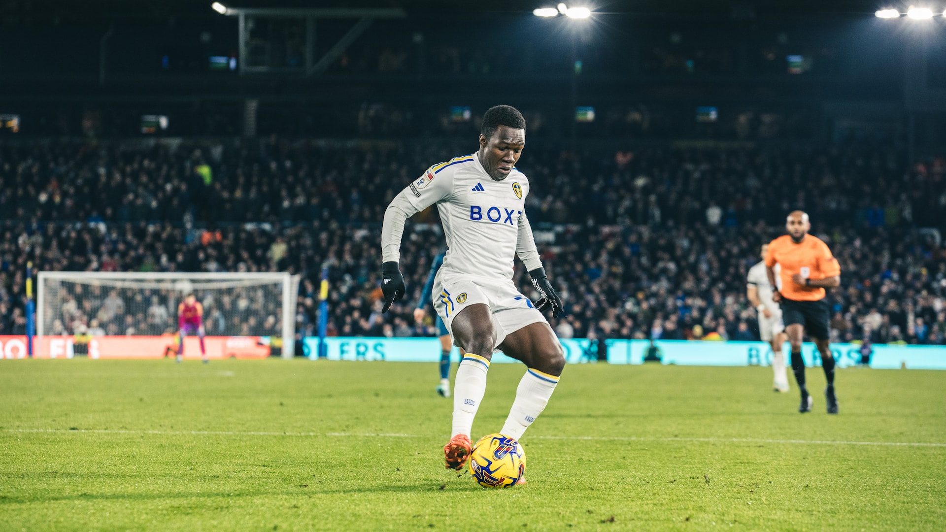 A photograph of Wilf Gnonto controlling a ball under the lights at Elland Road, wearing a long sleeve white home kit and gloves
