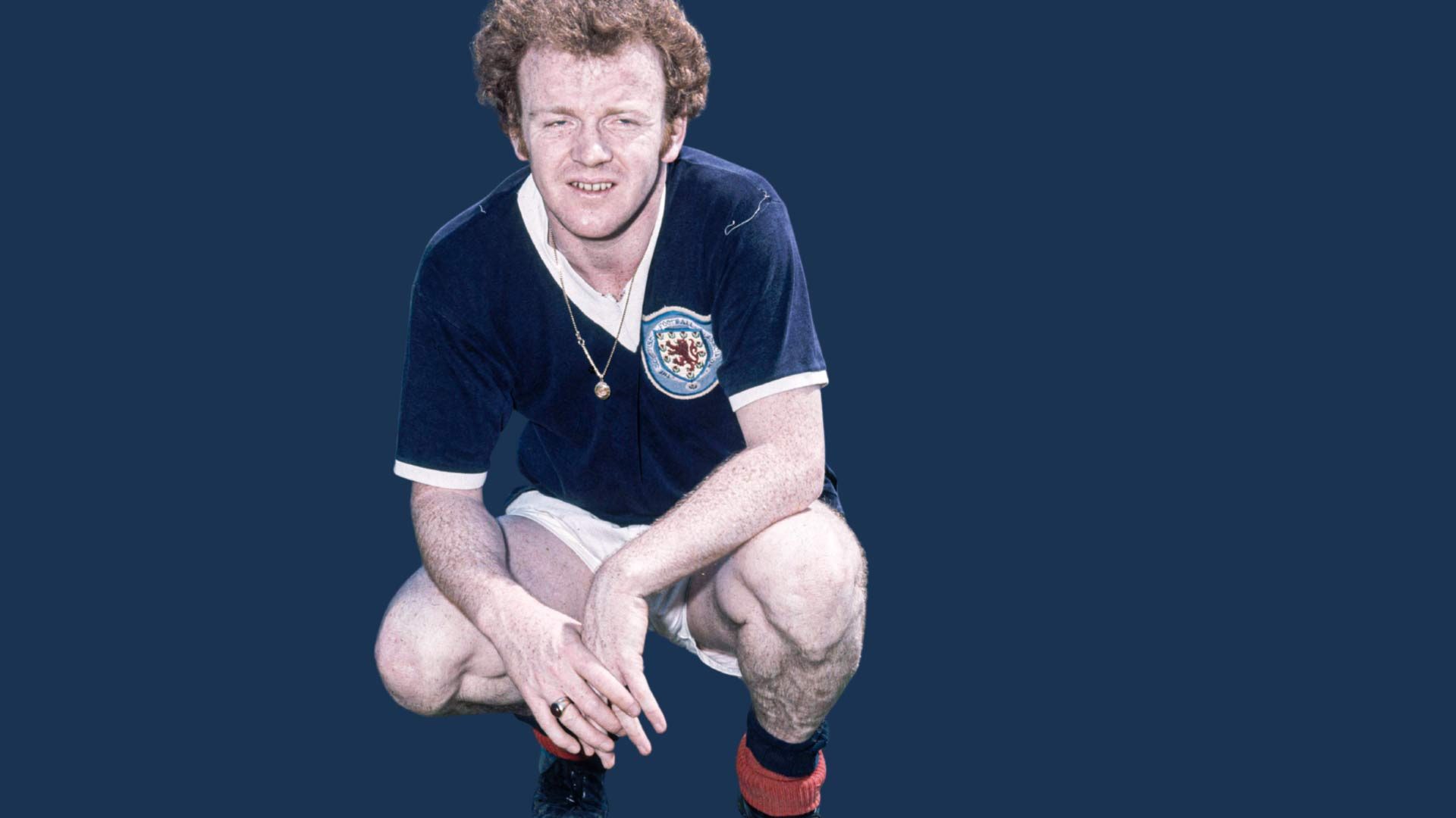 Billy Bremner kneeling down in a Scotland kit, necklace hanging out, looking absolutely haggard